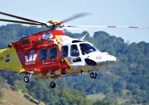 A red, yellow and white emergency rescue helicopter with the Westpac bank logo on the side hovers with some Australian bushland in the background.